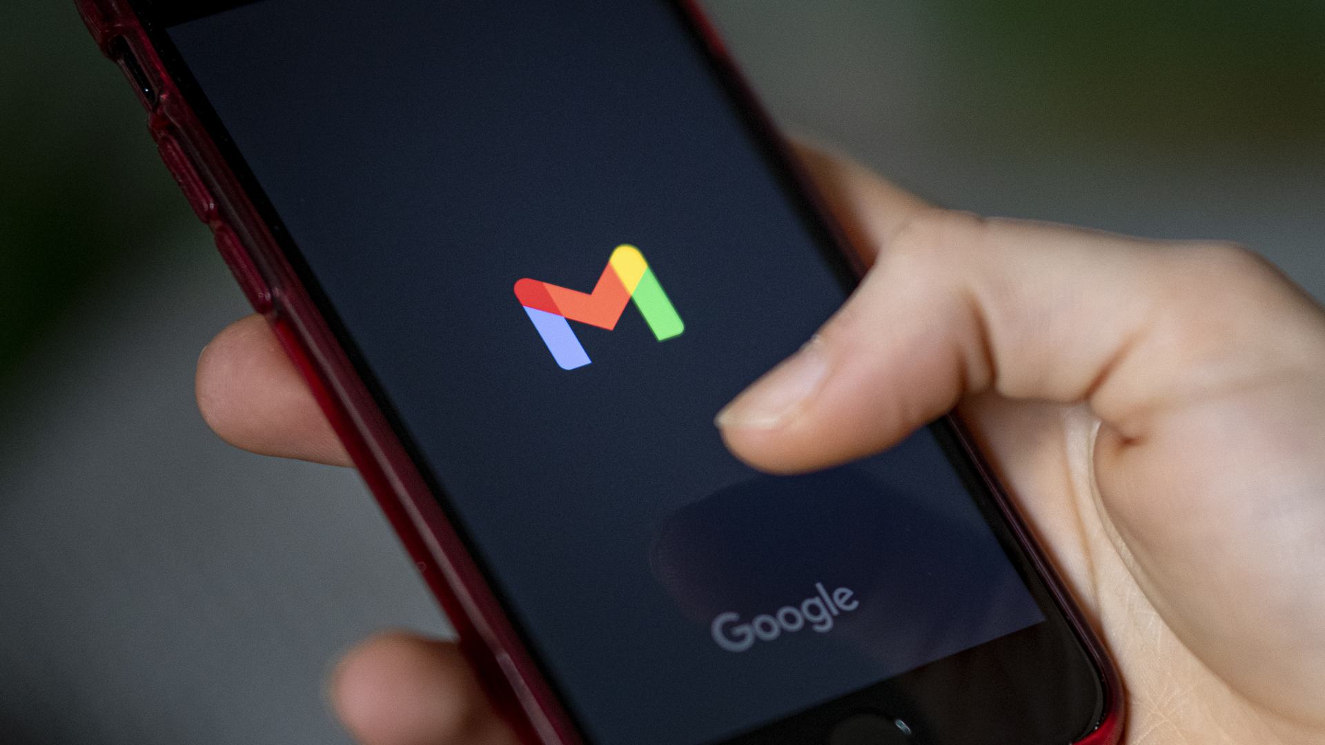 A hand holds a smartphone showing the logo of the Gmail app and the word “Google” on the screen.