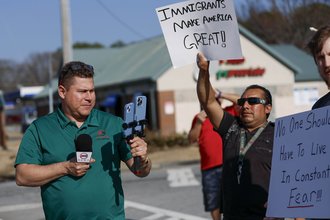 Mario Geuvara stands on a street holding a microphone and two cellphones, recording himself, beside protesters holding pro-immigration signs.