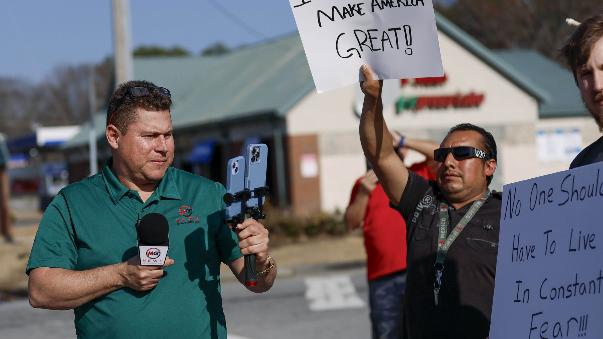 Mario Geuvara stands on a street holding a microphone and two cellphones, recording himself, beside protesters holding pro-immigration signs.
