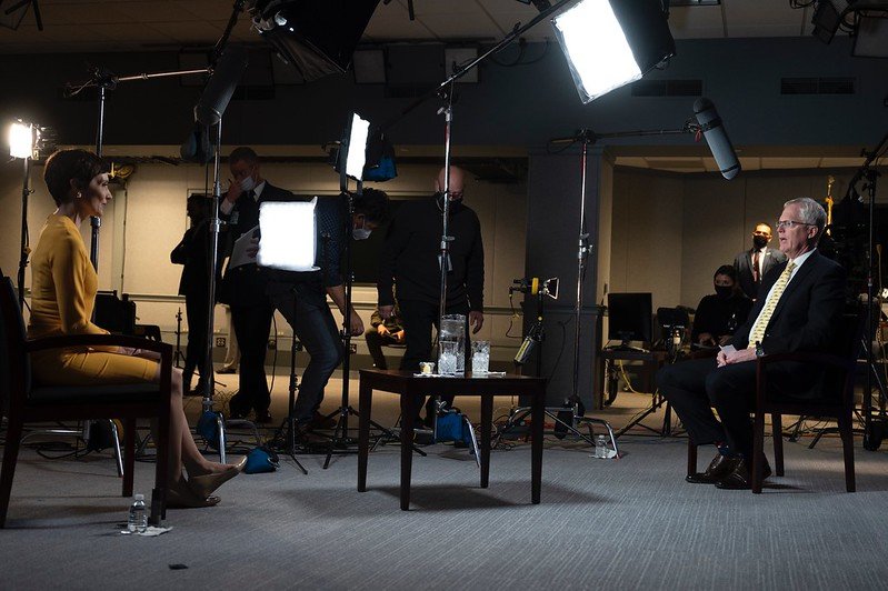 Reporter Catherine Herridge, left, wears a yellow dress and sits in front of lights and microphones, interviewing Acting Defense Secretary Chris Miller, who wears a dark suit and sits, right