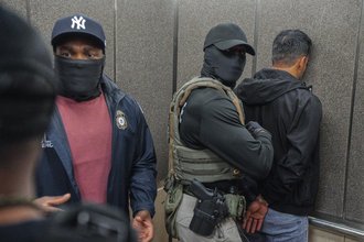 An ICE agent wearing a New York Yankees ballcap with a mask over his face showing only his eyes faces the camera while a second ICE agent wearing a black ballcap and mask that entirely obscures his face holds a handcuffed man face first against a wall.