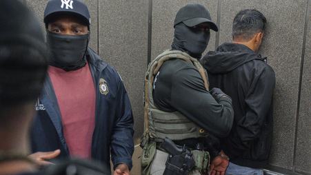An ICE agent wearing a New York Yankees ballcap with a mask over his face showing only his eyes faces the camera while a second ICE agent wearing a black ballcap and mask that entirely obscures his face holds a handcuffed man face first against a wall.