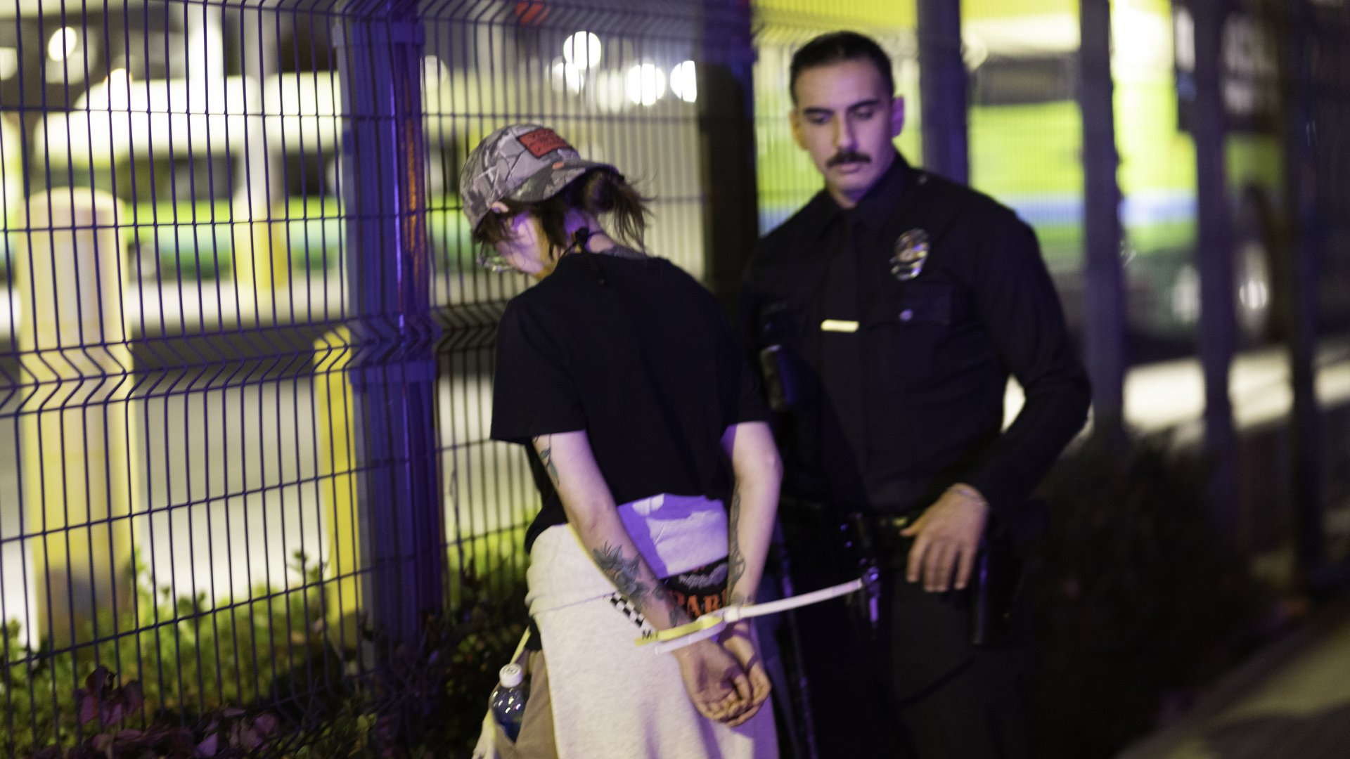 Mel Buer stands with her back to the camera, showing her hands zip-tied behind her back, while a police officer stands next to her, facing the camera.