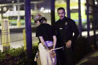 Mel Buer stands with her back to the camera, showing her hands zip-tied behind her back, while a police officer stands next to her, facing the camera.