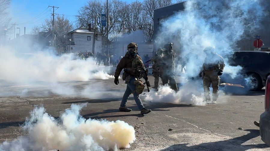 The image shows a street scene where several individuals in tactical gear are surrounded by thick white smoke filling the air. One individual in the foreground is walking across the street wearing a gas mask, helmet, and body armor, and carrying a rifle. Other figures in tactical gear can be seen through the smoke behind the person in the foreground. In the background, there are buildings and bare trees under a bright blue sky, with a crowd of people gathered further down the street.