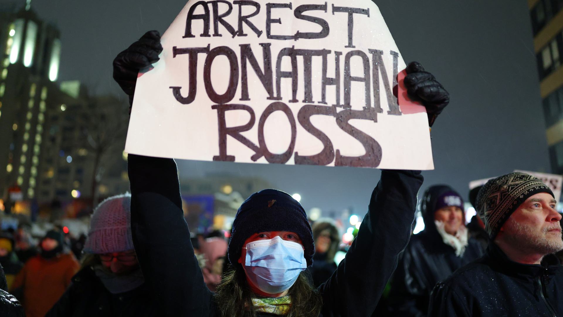 A protester wearing a medical mask and dark hooded shirt, shown at night in a crowd of other protesters, holds up a white sign with the following written in black “ARREST JONATHAN ROSS”