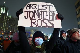 A protester wearing a medical mask and dark hooded shirt, shown at night in a crowd of other protesters, holds up a white sign with the following written in black “ARREST JONATHAN ROSS”