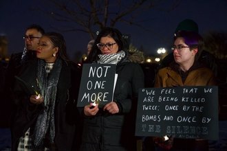 Rep. Cori Bush, Rep. Rashida Tlaib, and a vigil attendee stand outside. Rep. Bush holds a candle, and Rep. Tlaib holds a sign stating "Not One More"