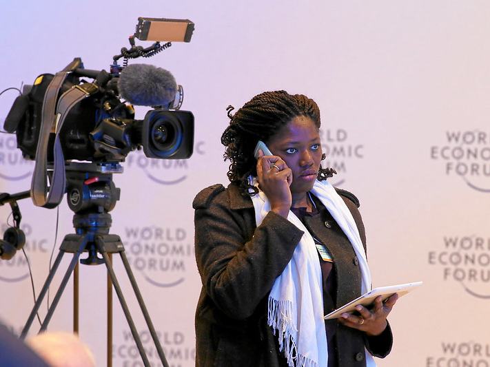 A journalist stands next to a television camera and talks on her mobile phone in front of a backdrop for the World Economic Forum.