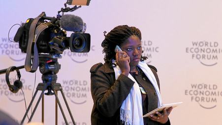 A journalist stands next to a television camera and talks on her mobile phone in front of a backdrop for the World Economic Forum.