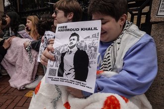 A person sits chained to a fence with their face partially obscured by a poster with a picture of Mahmoud Khalil and the words “Free Mahmoud Khalil”