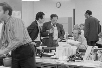 Black and white photograph focused on three men in a newsroom conferring. Two are standing and one is sitting at a desk, holding papers.