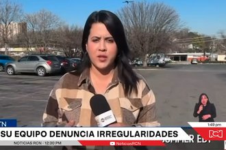 Screenshot of a news report showing a female reporter in a brown checked shirt and with long dark hair standing in a parking lot in daytime and speaking into a microphone, with a station chyron displaying text in Spanish.