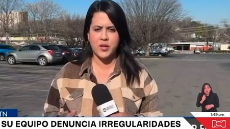 Screenshot of a news report showing a female reporter in a brown checked shirt and with long dark hair standing in a parking lot in daytime and speaking into a microphone, with a station chyron displaying text in Spanish.