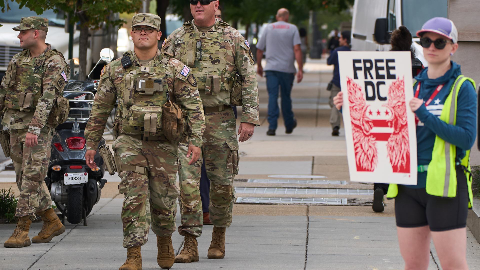 Three members of the National Guard walking on a street in daylight, as a protester with a sign saying “FREE DC” stands nearby.