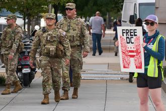 Three members of the National Guard walking on a street in daylight, as a protester with a sign saying “FREE DC” stands nearby.