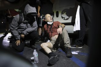 A man with camera equipment around his neck sits on the ground with one pant leg pulled up, showing a bandage around his thigh. Another person in a dark jacket with the hood pulled over their face kneels next to him.