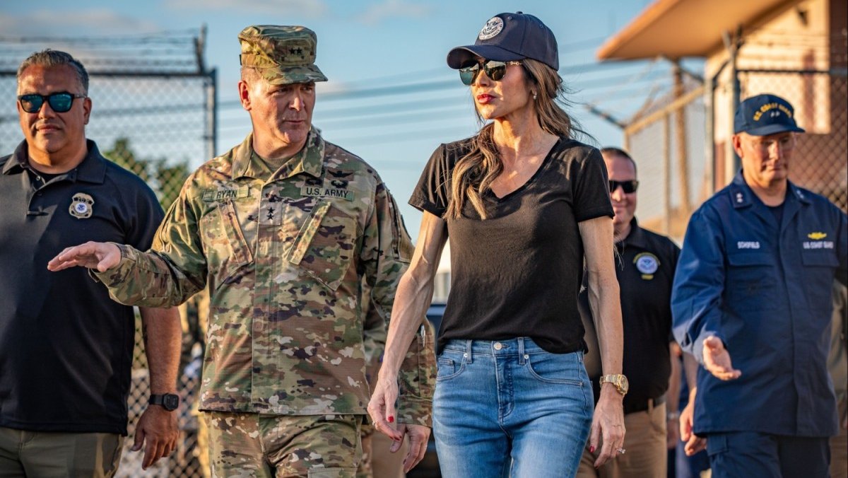 Kristi Noem, wearing jeans, a black T-shirt, sunglasses, and a baseball cap stands outside next to a U.S. Army member in fatigues and several other men on an official visit to Guantánamo Bay, Cuba.