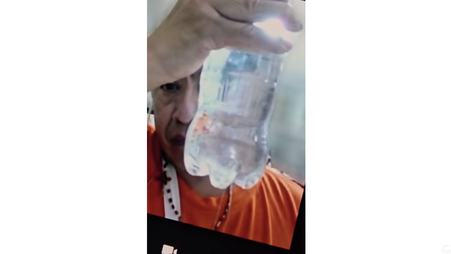 A screenshot of Carlitos Ricardo Parias in detention, holding up a bottle of water in which small wormlike creatures are floating.