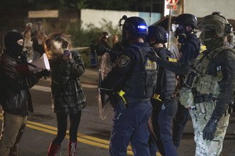Nighttime scene of four federal agents standing on a street across from two protesters. One agent holds up a shield and canister out of which he sprays brown liquid directly into the face of a female protester, who is standing and holding up a phone, as another protester holding a megaphone looks on.