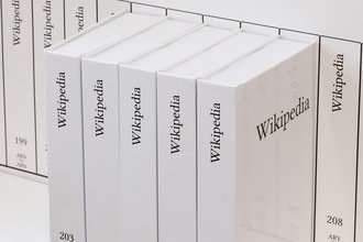 Five white books with “Wikipedia” written on their spines and the volume numbers 203, 204, 205, 206, and 207, respectively, sit on a white shelf in front of numerous other volumes of the printed Wikipedia.