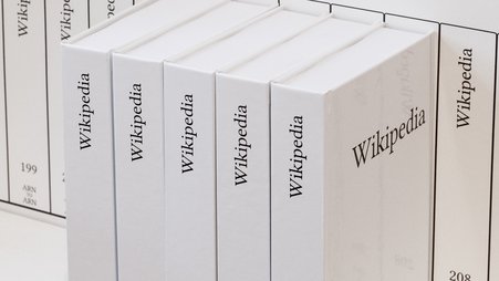Five white books with “Wikipedia” written on their spines and the volume numbers 203, 204, 205, 206, and 207, respectively, sit on a white shelf in front of numerous other volumes of the printed Wikipedia.