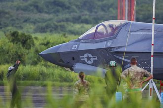 The nose and cockpit of a fighter jet on a runway surrounded by greenery, as two people in military fatigues look on. Two aircraft marshallers stand on the runway wearing white and green helmets, one with his arms raised.