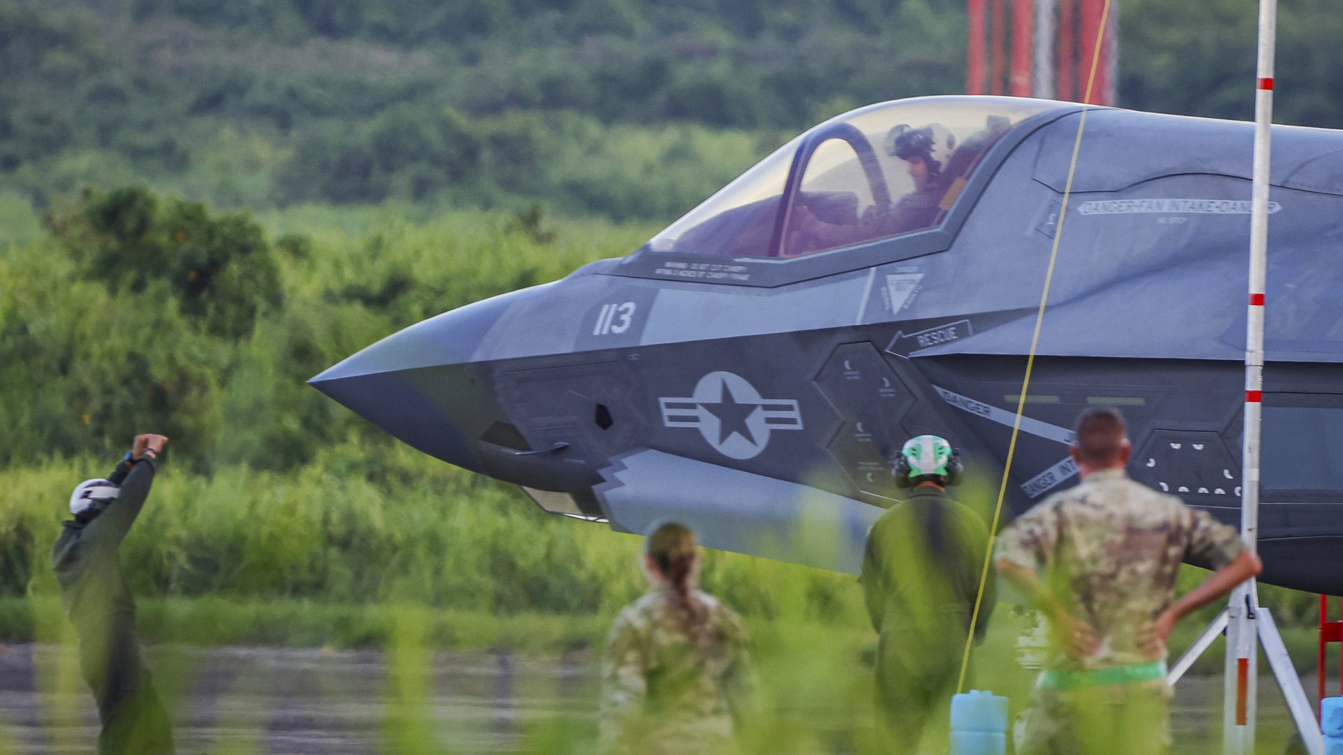 The nose and cockpit of a fighter jet on a runway surrounded by greenery, as two people in military fatigues look on. Two aircraft marshallers stand on the runway wearing white and green helmets, one with his arms raised.