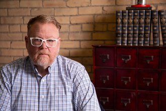 Bryan Schott, wearing a blue checkered shirt and white glasses, sits before a brick wall and dresser with books.