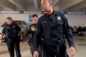 Three police officers stand, with one in the foreground and two in the background, in a parking garage. In the background on the right, several people can be seen sitting against a wall with their hands behind their backs.