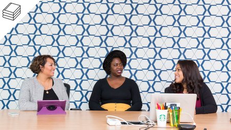Three women sitting at an office table in front of computers and tablets.