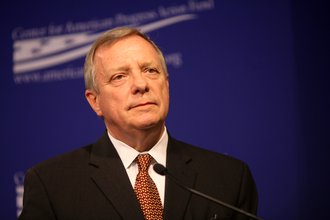 Senator Durbin, wearing a black suit and a red patterned tie, stands at a microphone in front of a blue background.