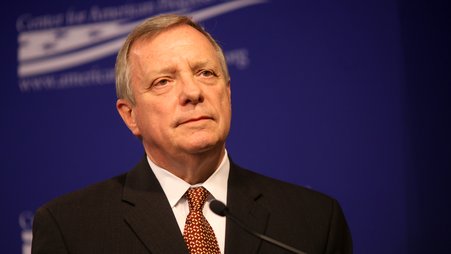 Senator Durbin, wearing a black suit and a red patterned tie, stands at a microphone in front of a blue background.