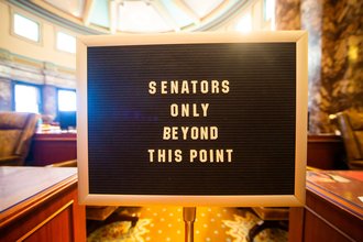 A letter board sign stands before a legislative chamber and reads "Senators only beyond this point"