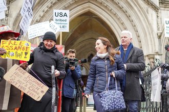 Stella Assange, wearing a blue jacket and holding a blue bag, smiles as she stands outside the UK high court, surrounded by protestors supporting Assange.