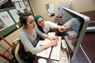 A woman in a striped shirt sits at a computer keyboard, typing, as another woman in a bluehat looks on and points at the screen.