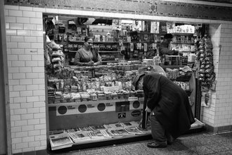 A man in a dark jacket and hat bends down to pick up a newspaper from a subway newsstand