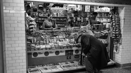A man in a dark jacket and hat bends down to pick up a newspaper from a subway newsstand