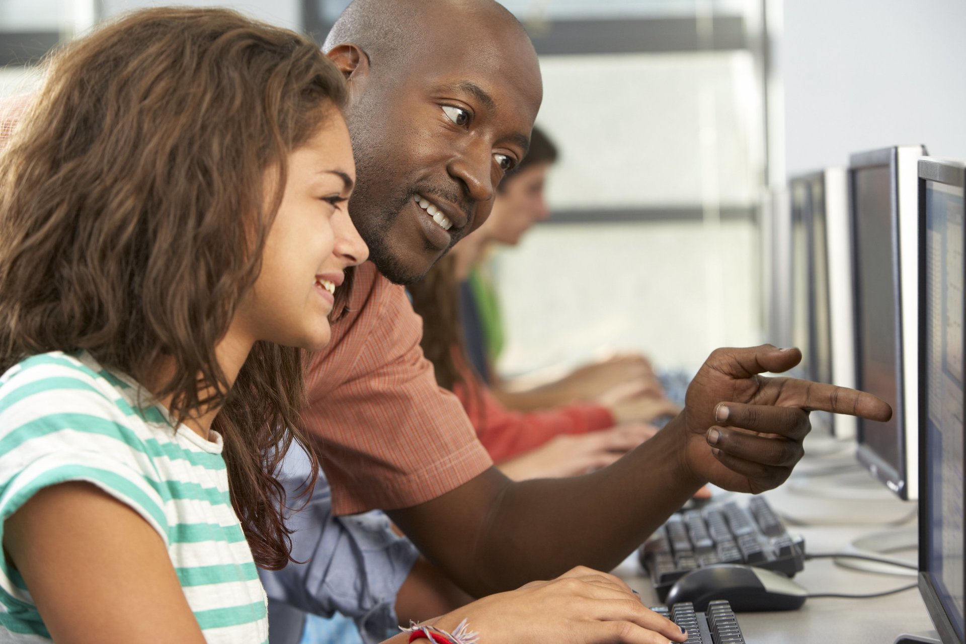 A smiling female student sits at a computer in the foreground, as a teacher leans over her and points to the screen in the background.