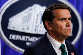 Todd Blanche, frowning and wearing a dark suit with an American flag pin, shown from the chest up in front of a Department of Justice seal