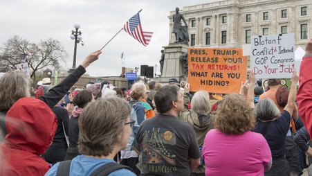 A crowd of protestors stand before a statue and building, with one holding an orange sign reading, "Pres. Trump: Release your tax returns! What are you hiding?"