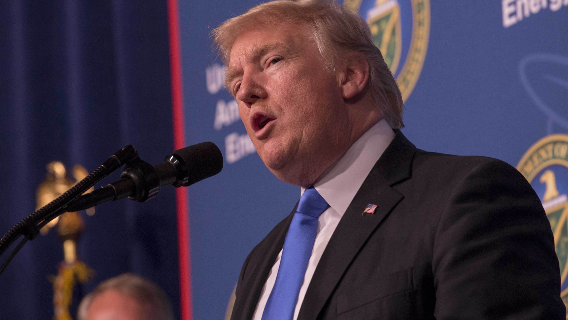 Former President Donald Trump, wearing a black suit jacket, white shirt, and blue tie, stands and speaks into a microphone