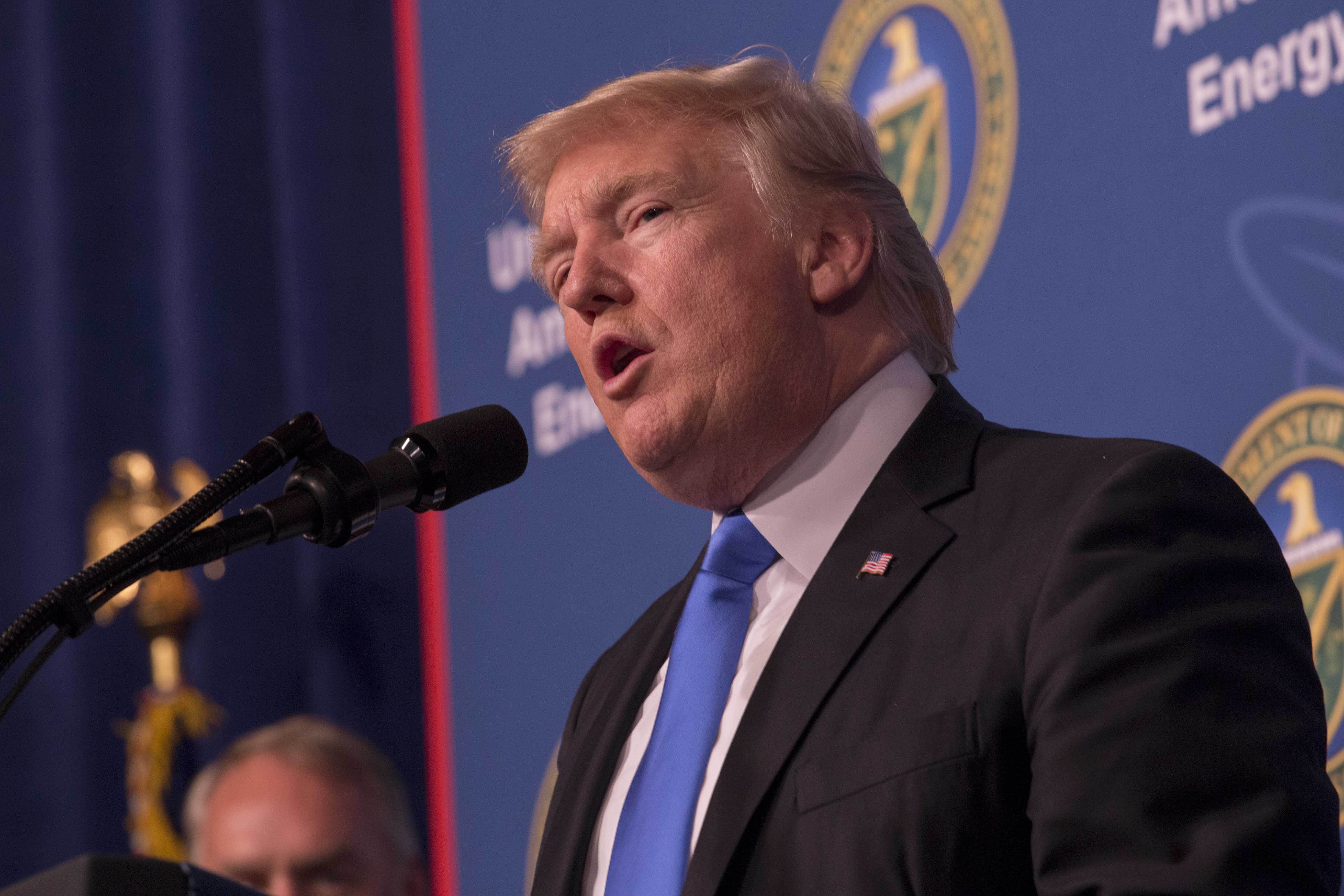 President Trump, wearing a black jacket and blue tie, speaks into a microphone