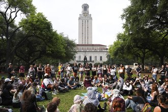 A crowd of people, some standing and some sitting on a grassy area surrounded by trees in front of The Main Tower, a tall tower at the University of Texas at Austin.