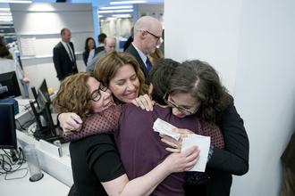 Four women hug in a brightly lit room, standing before computers and others milling around in the background.