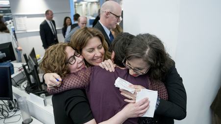 Four women hug in a brightly lit room, standing before computers and others milling around in the background.