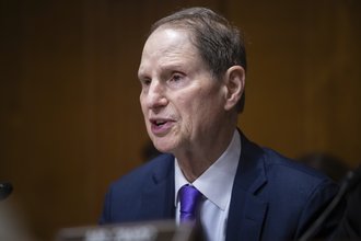 Sen. Ron Wyden, shown from the chest up, wearing a dark suit and sitting in a wood-paneled room, speaking into a microphone and pointing forward.