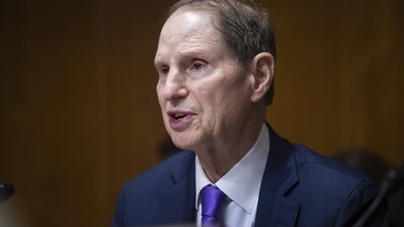 Sen. Ron Wyden, shown from the chest up, wearing a dark suit and sitting in a wood-paneled room, speaking into a microphone and pointing forward.