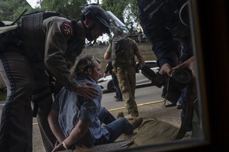 A man in a blue shirt sits, handcuffed, while a police officer wearing a helmet with a face shield lifted stands over him with his hands on the man's shoulders. The hands of another officer can be seen holding a camera.