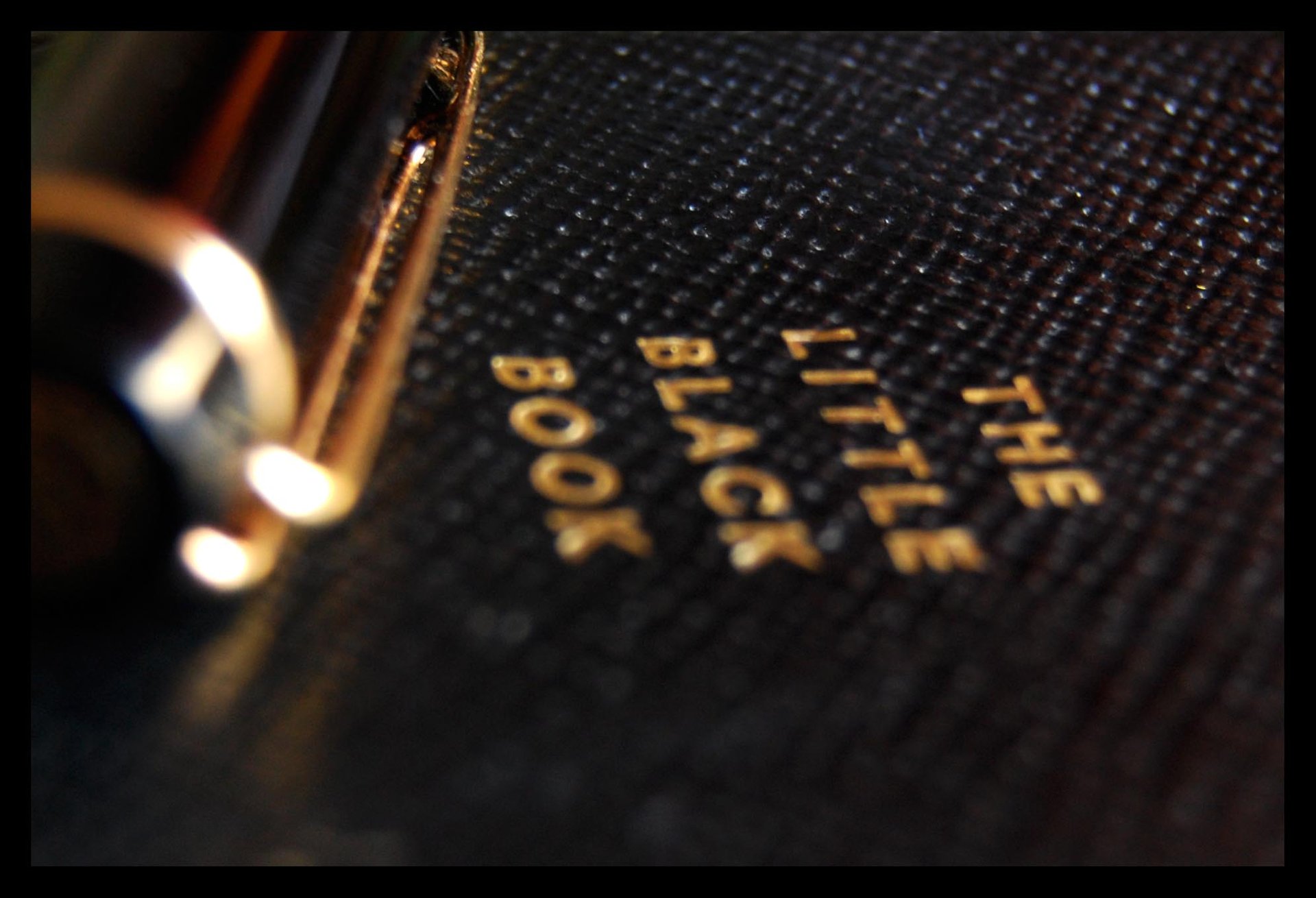 Macro lens photo of a black notebook titled "The little black book" in gold lettering with a black and gold pen on it